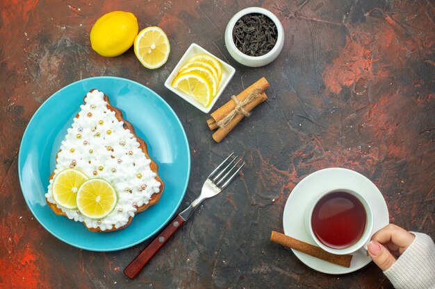 Top view cake with lemon on blue plate lemon slices in bowl fork cup of tea in female hand cinnamon sticks on dark red background