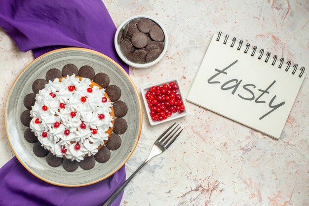 Top view cake with pastry cream and chocolate on plate purple shawl bowls with chocolate and berries fork tasty written on notebook