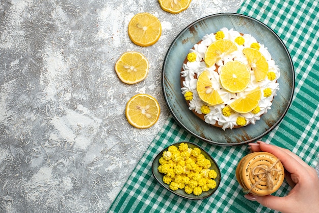 Top view cake with pastry cream and lemon on platter cookies in female hand candies in bowl on green white checkered tablecloth