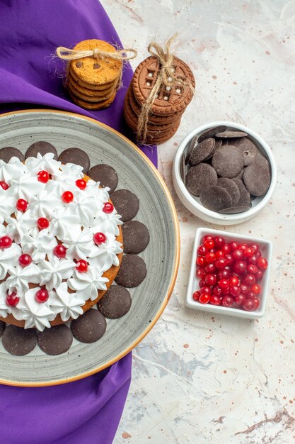 Top view cake with pastry cream on plate purple shawl cookies tied with rope chocolate and berries in bowls on white table