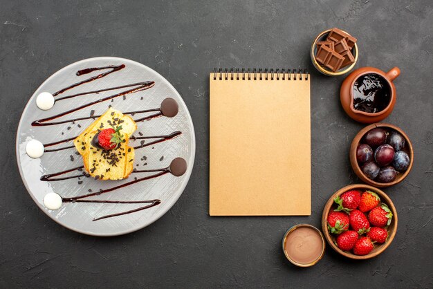 Top view cake with strawberry cream notebook between strawberries chocolate in bowls and plate of cake with chocolate sauce on the dark surface