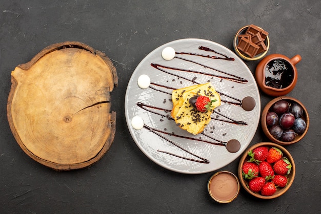 Top view cake with strawberry strawberries chocolate and berries in bowls and plate of cake with strawberries and chocolate sauce next to the cutting board on table