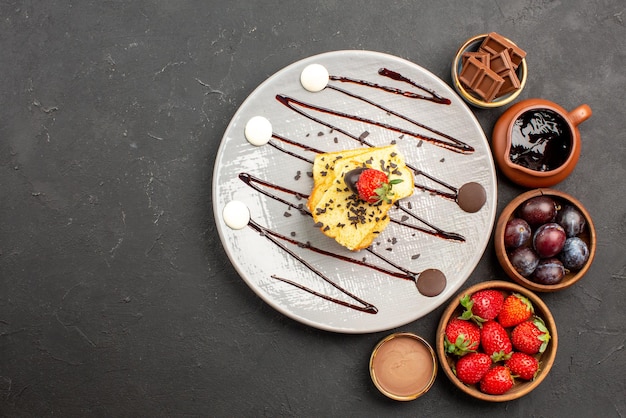 Top view cake with strawberry strawberries chocolate and berries in bowls and plate of cake with strawberries and chocolate sauce on table