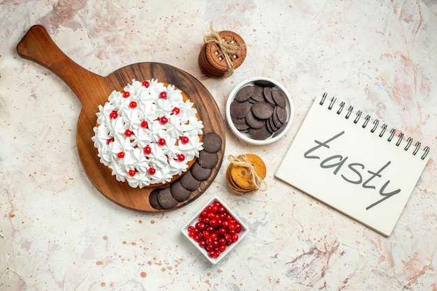 Free Photo top view cake with white pastry cream on chopping board bowls with berries and chocolate cookies tied with rope tasty written on notebook on light grey background