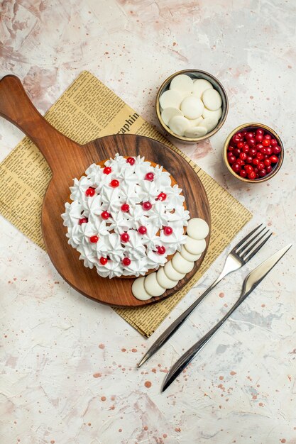 Top view cake with white pastry cream on wood board on newspaper fork and dinner knife berries and white chocolate in bowls