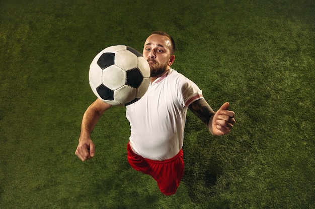 Top view of caucasian football or soccer player on green background of grass. Young male sportive model training, practicing. Kicking ball, attacking, catching. Concept of sport, competition, winning.