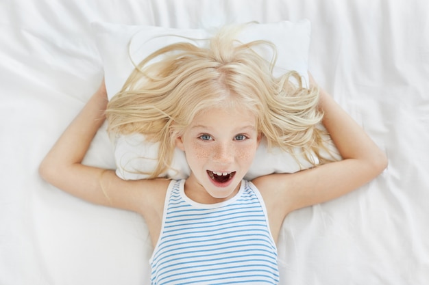 Free photo top view of cheerful little girl with blonde hair, feckles and blue eyes wearing stripped pajamas lying on white pillow and linen in her bed, having fun and laughing, doesn't want to have daytime nap