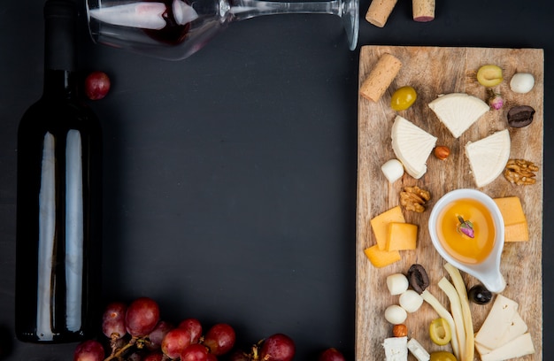 Top view of cheese set with cheddar brie string feta and butter olive walnuts on cutting board with bottle and glass of red wine and corks on black with copy space