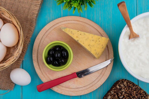 Free photo top view cheese with olives and a knife on a stand with yogurt in a bowl on a turquoise background