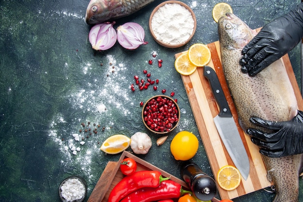 Top view of chef with black gloves holding raw fish on wood board pepper grinder flour bowl pomegranate seeds in bowl on kitchen table