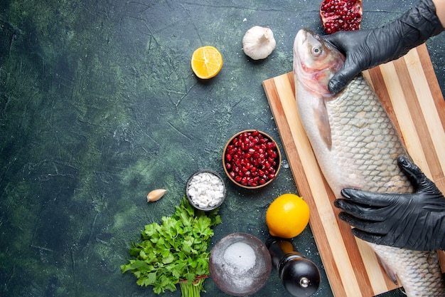 Free photo top view chef with black gloves holding raw fish on wood board pepper grinder pomegranate seeds in bowl on table free space