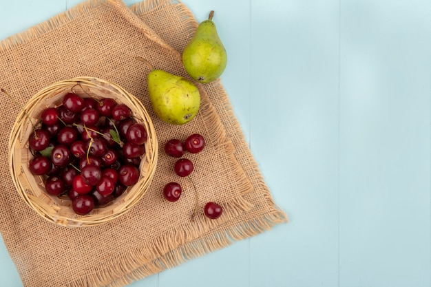 Top view of cherries in basket and pears on sackcloth on blue background with copy space