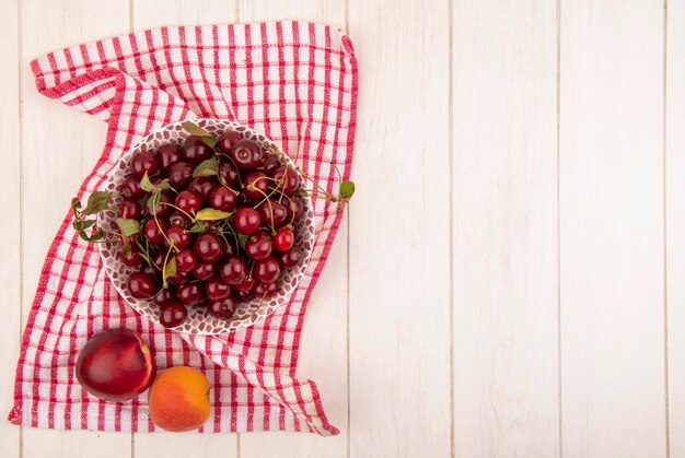 Top view of cherries in bol and peaches on plaid cloth and wooden background with copy space