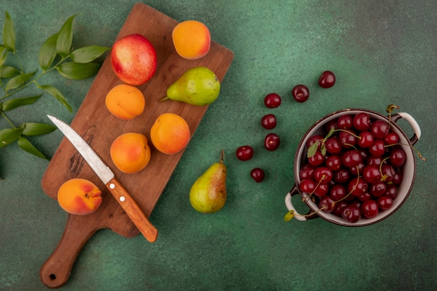 Top view of cherries in bowl and pattern of apricots pear peach with knife on cutting board and leaves on green background