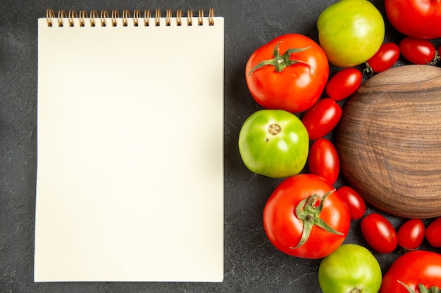 Top view cherry red and green tomatoes around a wooden plate and a notebook on dark ground