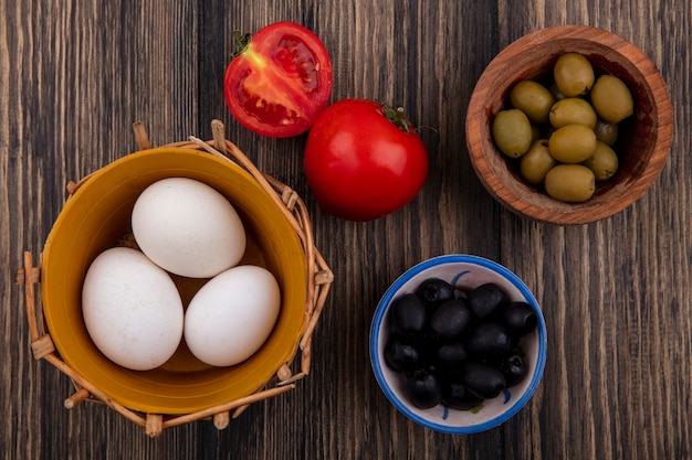 Top view chicken eggs in basket with black and green olives in bowls and tomatoes on wooden background