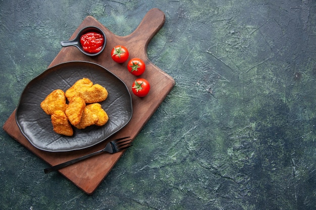 Top view of chicken nuggets on a black plate and fork on wooden board tomatoes ketchup on the right side on dark surface