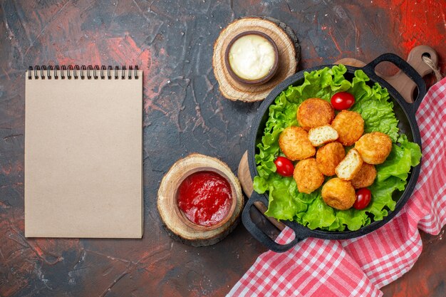 Top view chicken nuggets cherry tomatoes lettuce in pan sauce bowls on wood boards notebook on dark red wall