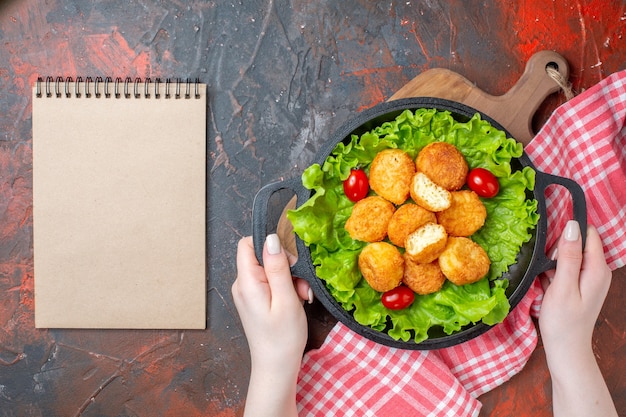 Top view chicken nuggets lettuce cherry tomatoes in pan in female hands notebook on dark red wall