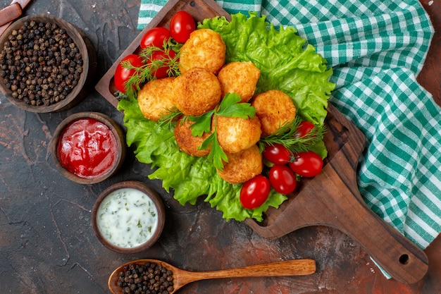 Top view chicken nuggets lettuce cherry tomatoes on wood board black pepper in bowl sauces in small wooden bowls wooden spoon on dark surface