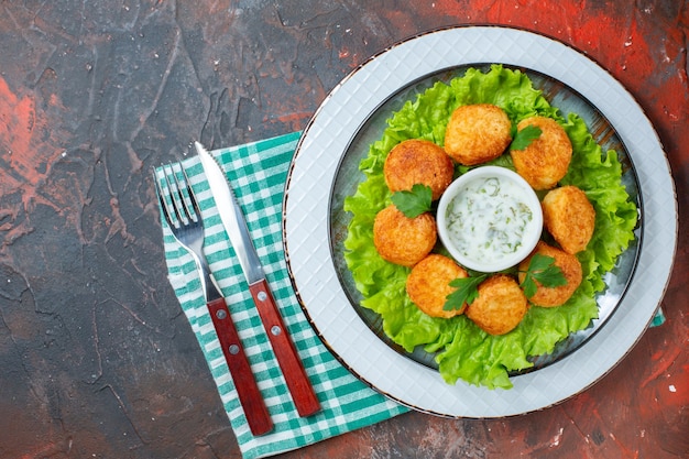 Top view chicken nuggets lettuce and sauce on plate fork and knife on dark table