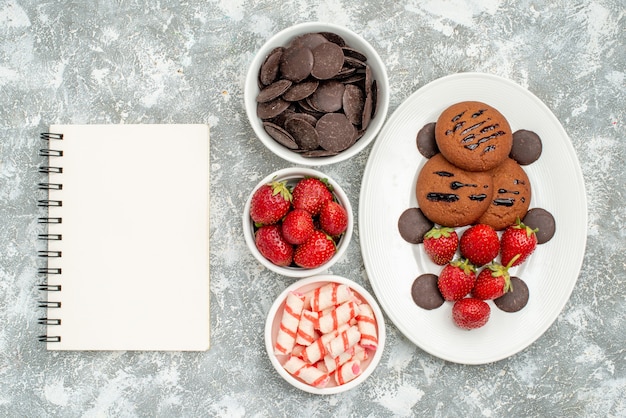 Top view chocolate cookies strawberries and round chocolates on the white oval plate and bowls with candies strawberries chocolates and a notebook on the grey-white ground