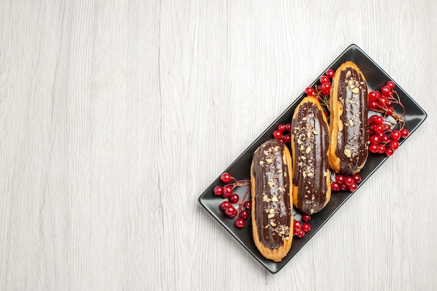 Free photo top view chocolate eclairs and currants on the black isometric rectangular plate at the right side of the white wooden ground