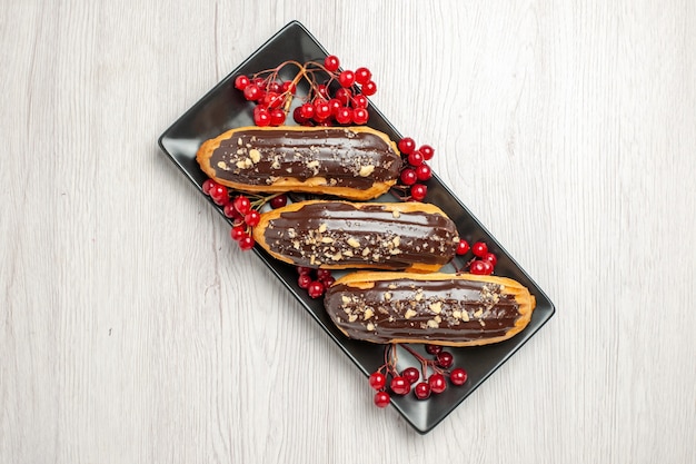 Top view chocolate eclairs and currants on the black rectangular plate at the center of the white wooden table