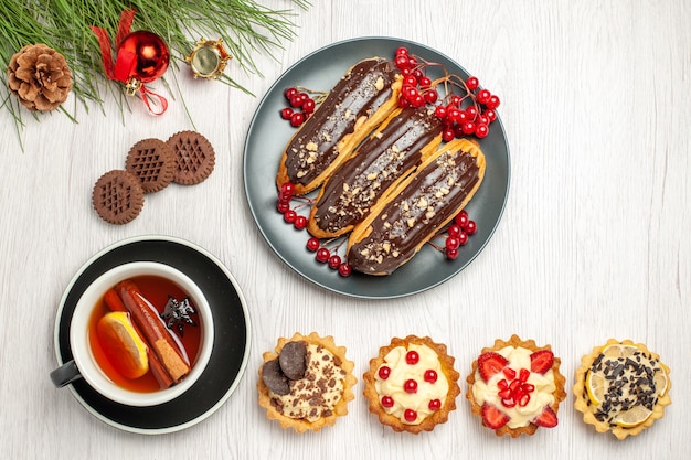 Free photo top view chocolate eclairs and currants on the grey plate lemon cinnamon tea tarts cookies and pine tree leaves with christmas toys on the white wooden ground