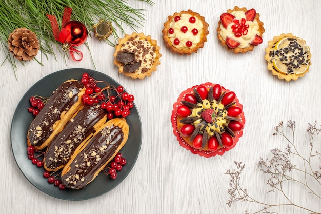 Top view chocolate eclairs and currants on the grey plate tarts berry cake and pine tree leaves with christmas toys on the white wooden table