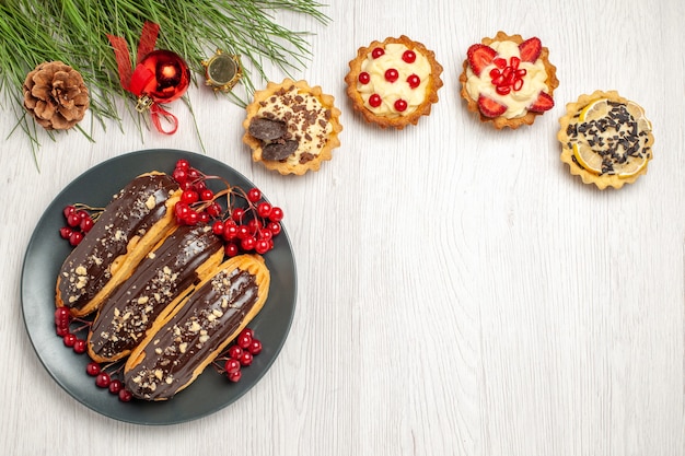 Top view chocolate eclairs and currants on the grey plate tarts and pine tree leaves with christmas toys on the white wooden table