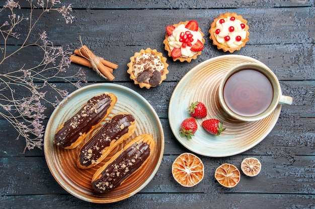 Top view chocolate eclairs on oval plate a cup o tea and strawberries on saucer tarts cinnamon and dried lemons on the dark wooden table