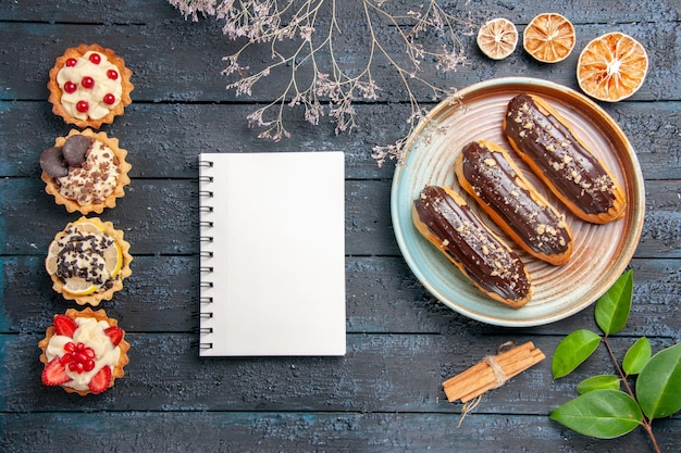 Free Photo top view chocolate eclairs on oval plate dried flower branch cinnamon dried oranges leaves a notebook and vertical row tarts on the dark wooden ground