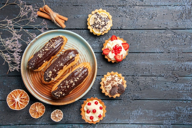 Free photo top view chocolate eclairs on oval plate surrounded with dried lemons and tarts at the left side of the dark wooden table with copy space