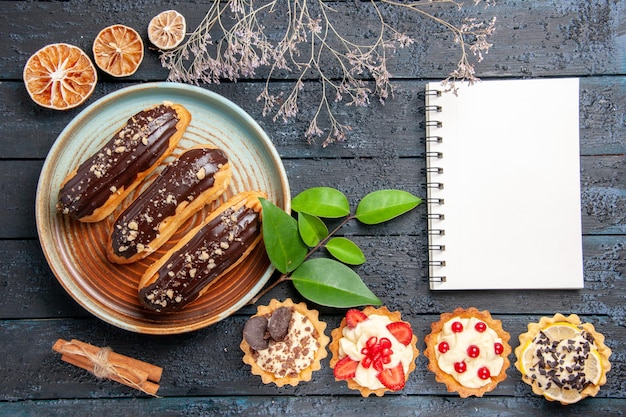 Free Photo top view chocolate eclairs on oval plate tarts laeves cinnamon dried oranges and a notebook on the dark wooden table