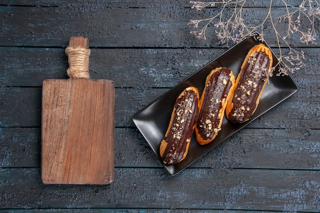 Free photo top view chocolate eclairs on rectangle plate and a chopping board on the dark wooden table with free space