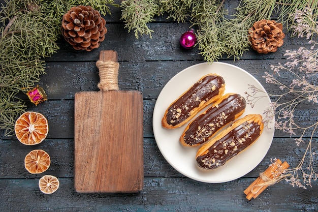 Top view chocolate eclairs on white oval plate fir-tree branches and cones christmas toys dried flower branch dried oranges cinnamon and cutting board on dark wooden table