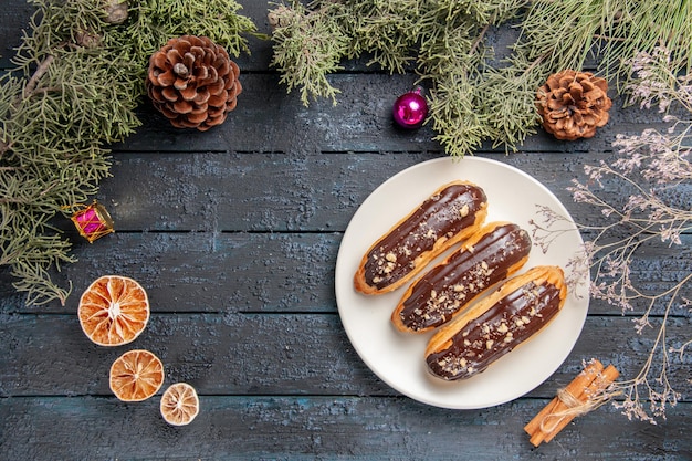 Top view chocolate eclairs on white oval plate fir-tree branches and cones christmas toys dried oranges cinnamon on dark wooden table with free space