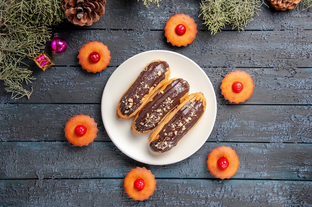 Top view chocolate eclairs on white oval plate surrounded with cupcakes fir-tree branches christmas toys on dark wooden ground with free space