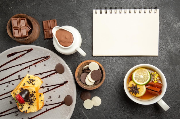 Top view chocolate on the table cake with strawberries and chocolate next to the cup of herbal tea and white notebook bowls of chocolate on the dark table