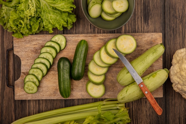 Free photo top view of chopped cucumbers and zucchinis isolated on a wooden kitchen board with knife with lettuce celery and cauliflower isolated on a wooden wall