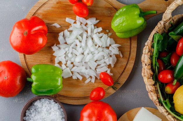 Top view chopped onions on cutting board with tomatoes, salt, green pepper on gray surface