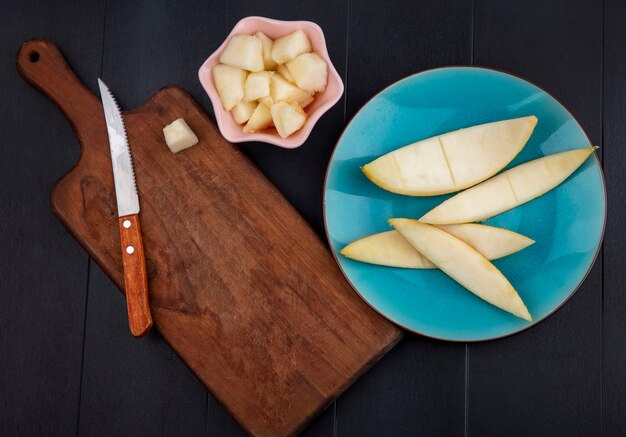 Top view of chopped slices of melon on pink bowl with peels on blue plate with wooden kitchen board with knife on black