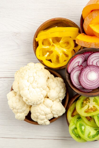 Top view chopped vegetables cut pumpkin cut yellow bell peppers cut onion cut green tomatoes cauliflower in bowls on white table