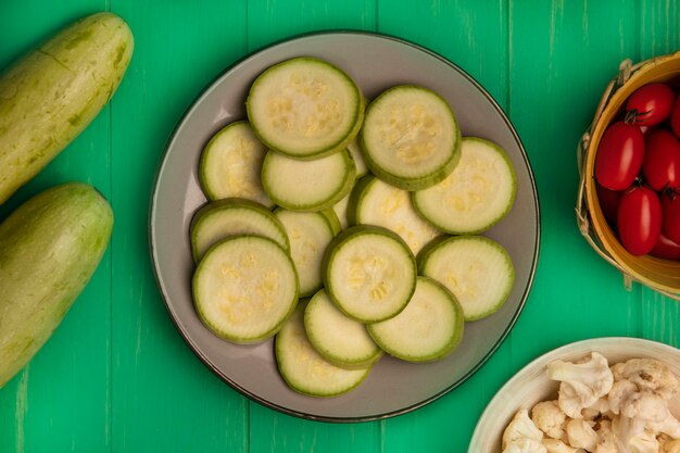 Top view of chopped zucchini slices on a plate with plum tomatoes on a bucket with cauliflower florets on a bowl with zucchinis isolated on a green wooden wall