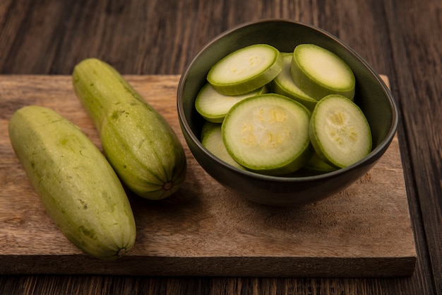 Top view of chopped zucchinis on a bowl with whole zucchinis isolated on a wooden kitchen board on a wooden wall