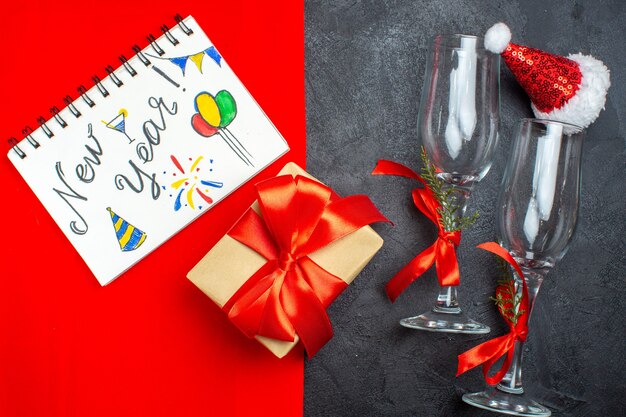 Top view of Christmas background with santa claus hat glass goblets notebook with new year writing and drawings and gift on red and black background