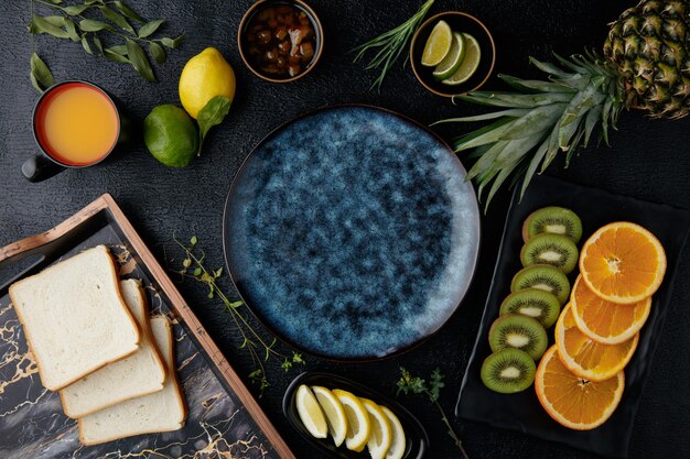 Top view of citrus fruits as pineapple kiwi orange lime lemon and tangerine slices with bread slices in tray and jam in bowl around empty plate on black background