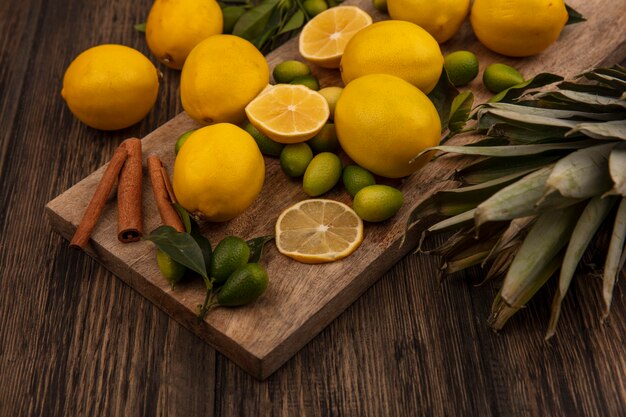 Top view of citrus fruits such as kinkans and lemons with cinnamon sticks on a wooden kitchen board with pineapples on a wooden background