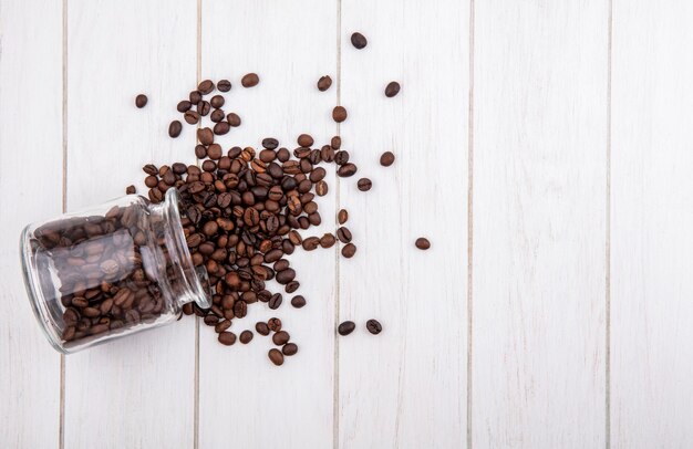 Top view of coffee beans falling out of a glass jar on a white wooden background with copy space